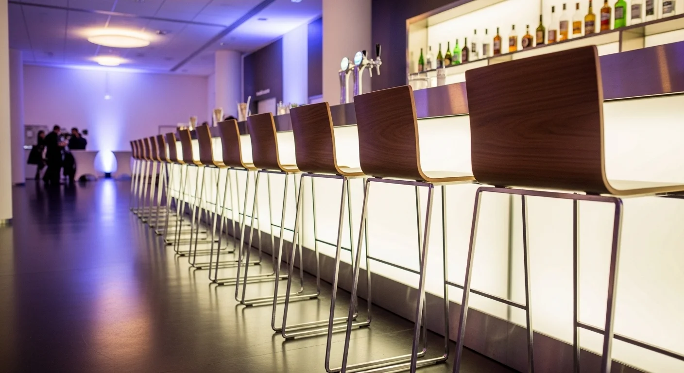 Fila de bancos altos de diseño metálico y madera en un evento corporativo, frente a una barra iluminada.